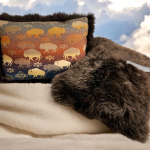 Peut inclure: Deux coussins décoratifs, l'un avec une housse en fausse fourrure brune et l'autre avec un tissu à motifs colorés représentant un motif répété de bisons stylisés. Le coussin à motifs a une bordure brun foncé.