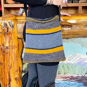 Può includere: Una borsa a tracolla con un motivo a righe grigie, gialle e blu navy. La borsa è rettangolare e ha una tracolla nera. La persona indossa un maglione nero a coste, leggings grigi e stivali da cowboy neri.