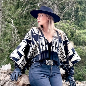 May include: A woman wearing a black hat, a black and white patterned poncho with fringe, a black belt with a decorative buckle, dark blue gloves, and blue jeans. The background features a forest setting with green trees.