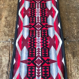 Puede incluir: Alfombra rectangular con un patrón geométrico en rojo, negro, gris y blanco. La alfombra tiene un borde negro y se muestra sobre una superficie de madera. El diseño presenta formas de diamante y flecha.
