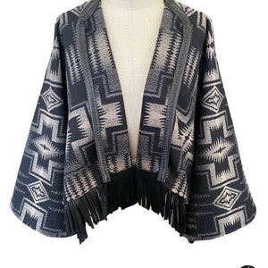 May include: A black and beige patterned jacket with a leather trim and fringe. The open-front jacket features a geometric design and two buttons. The fringe is along the bottom hem. The jacket is displayed on a mannequin.