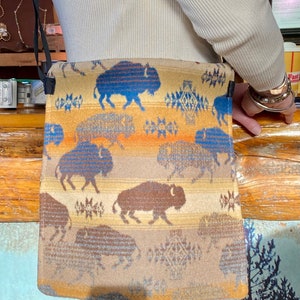 Pode incluir: Uma bolsa de ombro bege e marrom com um padrão de búfalo azul e marrom. A bolsa tem uma alça longa e um fecho de aba.