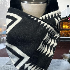 May include: Black and white geometric patterned neck warmer with a black fleece lining. The neck warmer is folded in half and has two black buttons.