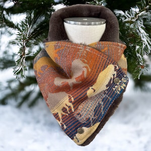 May include: A patterned scarf with a faux fur lining and snap closures. The scarf features a design with orange, brown, and blue tones, with animal silhouettes. The scarf is displayed against a snowy background with evergreen branches.