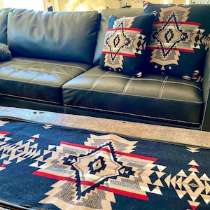 Puede incluir: Un sofá de cuero negro con tres cojines decorativos que presentan un patrón geométrico en tonos marrones, rojos y blancos. El sofá está delante de una mesa de café de madera con una manta de tiro de patrón azul y marrón.