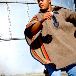 May include: A person wearing a brown poncho with colorful stripes and blue jeans. The poncho has a dark blue trim and a decorative buckle.