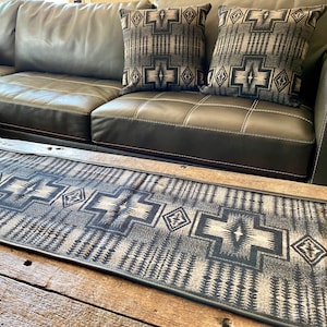 Puede incluir: Un sofá de cuero negro con dos almohadas de estampado blanco y negro. El sofá está delante de una mesa de café de madera con un corredor de estampado blanco y negro.