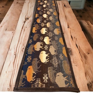 May include: A brown, blue and gold table runner with a repeating pattern of stylised buffalo silhouettes. The runner has a brown leather trim.
