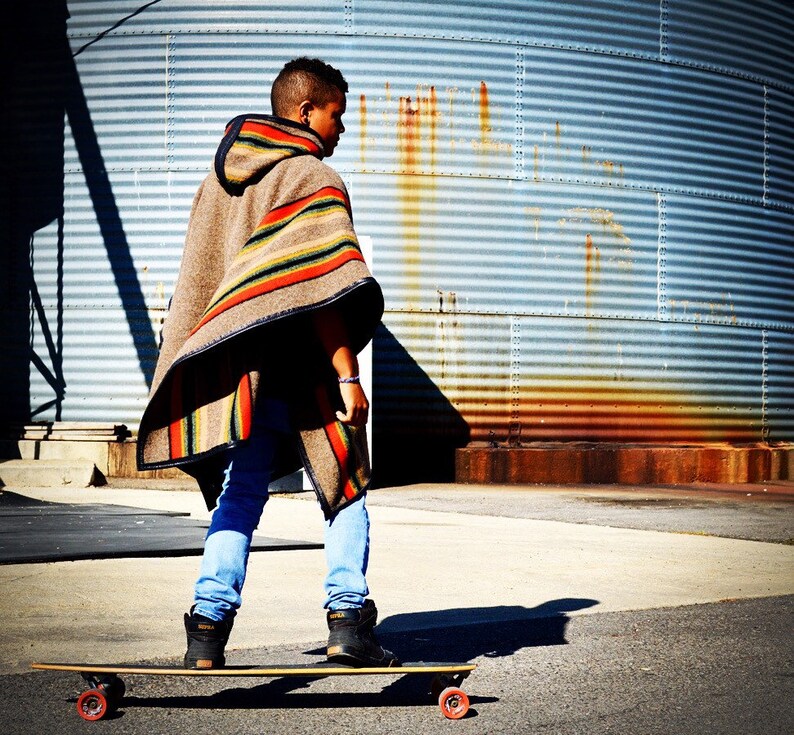 May include: A person wearing a brown and tan striped poncho with a hood, blue jeans, and black sneakers, standing on a longboard skateboard with black wheels.
