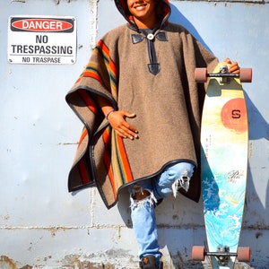 May include: A person wearing a brown hooded poncho with colorful stripes and blue jeans is standing next to a longboard with a colorful design. The longboard has black wheels and a red sun graphic on the deck. A sign that reads "DANGER NO TRESPASSING NO TRASPASE" is visible in the background.