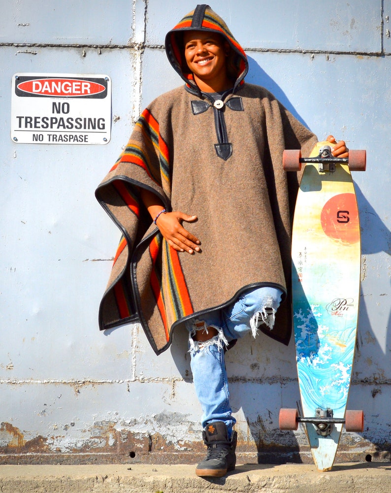 May include: A person wearing a brown hooded poncho with colorful stripes and blue jeans is standing next to a longboard with a colorful design. The longboard has black wheels and a red sun graphic on the deck. A sign that reads "DANGER NO TRESPASSING NO TRASPASE" is visible in the background.