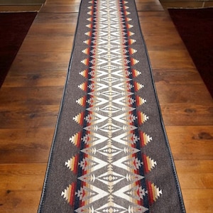 以下が含まれることがあります： 茶色、白、赤、オレンジ、青の色合いで幾何学模様が施された、長方形のテーブルランナー。ランナーは黒い縁取りがされている。