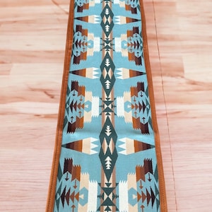 May include: A long, narrow table runner with a blue background and a geometric pattern in shades of brown, beige, and green. The runner has a brown leather border.