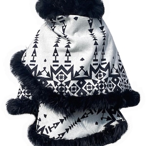 May include: A white and black patterned poncho with black faux fur trim. The poncho features a geometric design in black against a white background. The trim is thick and fluffy, adding a touch of luxury.