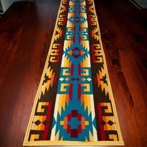 May include: A Southwestern-style table runner with a geometric pattern in shades of brown, blue, orange, yellow, and red. The runner has a yellow border.
