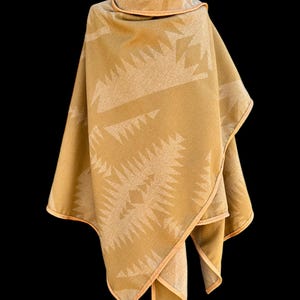 May include: A tan-colored poncho with a geometric pattern in a lighter shade of tan. The poncho has a brown leather trim around the edges.