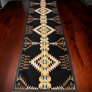 Puede incluir: Un camino de mesa negro con un patrón geométrico en tonos de marrón, naranja, amarillo y azul. El camino de mesa tiene un borde negro.