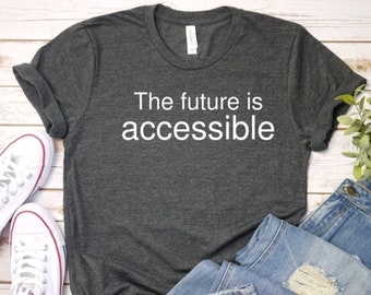 Może przedstawiać: Szary t-shirt z nadrukiem "The future is accessible" w kolorze białym na przodzie. T-shirt leży płasko na białym drewnianym tle z parą białych butów sportowych i niebieskimi dżinsami.