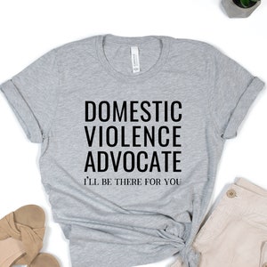 Op de afbeelding: Grijs T-shirt met de tekst "Domestic Violence Advocate" en "I'll be there for you" in zwart.