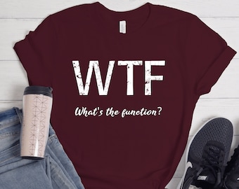 May include: Burgundy t-shirt with white text that reads "WTF What's the function?"  The shirt is laid flat on a white wooden surface with a pair of black sneakers, a pair of blue jeans, and a pink travel mug.