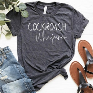 May include: A dark gray t-shirt with the text "COCKROACH Whisperer" in white lettering.