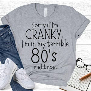 May include: A heather gray t-shirt with the text "Sorry if I'm CRANKY. I'm in my terrible 80's right now." The shirt is paired with blue jeans and white sneakers. A pair of glasses and a sudoku puzzle are also visible.