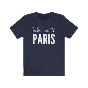 May include: A navy blue short-sleeved t-shirt featuring the white printed text "Take me to PARIS" centered on the chest. The design combines a cursive script font for the words "Take me to" positioned above the bold, capitalized word "PARIS".