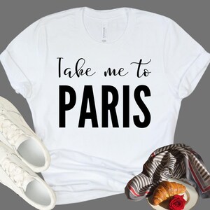 May include: A white short-sleeved t-shirt featuring the black text Take me to PARIS printed on the front. The shirt is displayed alongside a pair of white sneakers and a striped scarf with a croissant and a red rose placed on a small plate.