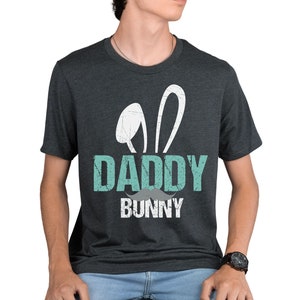 May include: A dark charcoal heather gray short-sleeve t-shirt featuring a graphic print with white bunny ears, the word DADDY in light teal, a gray mustache icon, and the word BUNNY in white. The design has a distressed, weathered texture.