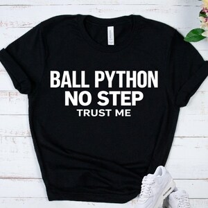 May include: Black t-shirt with white text that reads "BALL PYTHON NO STEP TRUST ME." The shirt has short sleeves and a crew neck. White sneakers are visible in the bottom right corner.