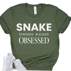 May include: An olive green short-sleeve t-shirt featuring the white text SNAKE STRESSED BLESSED OBSESSED printed on the front. The shirt has a crew neckline and a white brand label at the inner collar. A pair of white athletic sneakers is visible.
