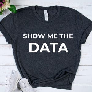 May include: Dark gray t-shirt with the text "SHOW ME THE DATA" in white block letters. The shirt is made of a soft material and has a classic crew neck design. White sneakers are visible in the bottom left corner.