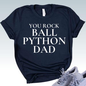 May include: Navy blue t-shirt with the white text "YOU ROCK BALL PYTHON DAD". The shirt has short sleeves and a crew neck. The shirt is paired with gray athletic shoes.