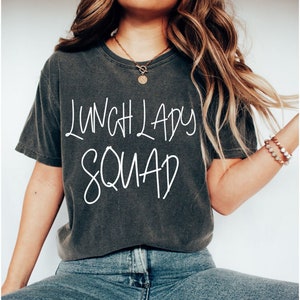 May include: A dark gray t-shirt with the text "Lunch Lady Squad" printed in white. The shirt is worn by a person with long brown hair and a gold necklace.