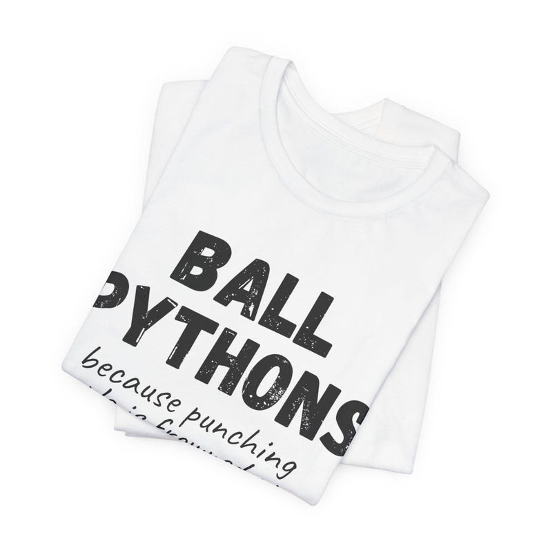 May include: White t-shirts with the text "BALL PYTHONS" in large, black, distressed font. The text "because punching..." is below. The shirts are folded and stacked on top of each other.