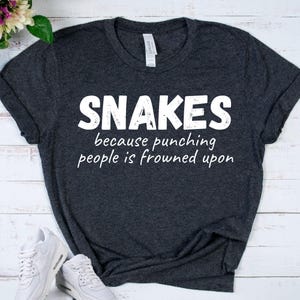 May include: A dark heather gray short-sleeve t-shirt with white text printed on the front that reads SNAKES because punching people is frowned upon. The shirt features a crew neckline and a standard fit with a visible brand label at the neck.