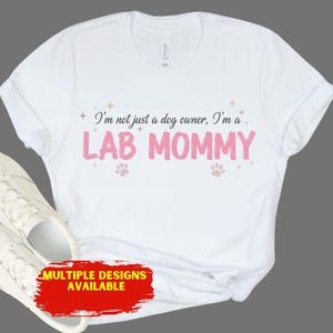 May include: White t-shirt with the text "I'm not just a dog owner, I'm a LAB MOMMY" in pink. The shirt also features paw prints and the text "MULTIPLE DESIGNS AVAILABLE" in a red box.