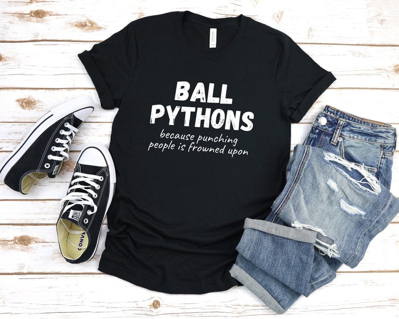May include: Black t-shirt with the text "BALL PYTHONS because punching people is frowned upon." The shirt is paired with blue jeans and black canvas sneakers with white laces.
