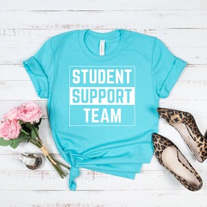 Może przedstawiać: Turkusowa koszulka z nadrukiem "STUDENT SUPPORT TEAM" w kolorze białym.