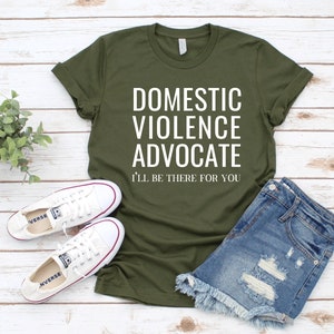 Op de afbeelding: Een groen T-shirt met de tekst "Domestic Violence Advocate I'll be there for you" in witte letters. Het shirt wordt gecombineerd met een spijkerbroekje en witte sneakers.