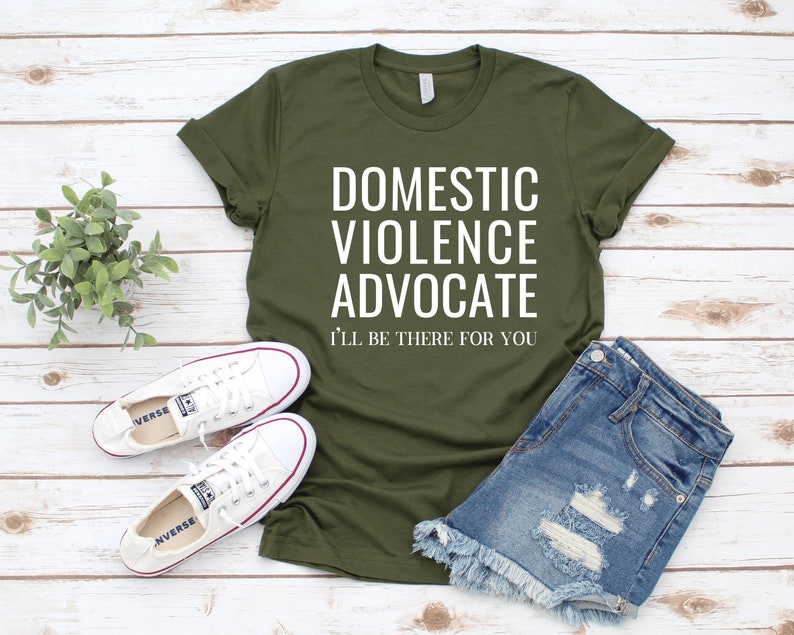 Op de afbeelding: Een groen T-shirt met de tekst "Domestic Violence Advocate I'll be there for you" in witte letters. Het shirt wordt gecombineerd met een spijkerbroekje en witte sneakers.