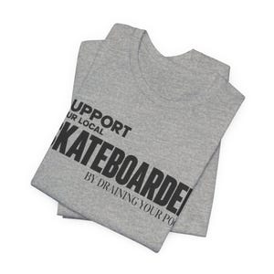 以下が含まれることがあります： SUPPORT YOUR LOCAL SKATEBOARDER BY DRAINING YOUR POOLという黒いテキストがプリントされたグレーのヘザーTシャツ。シャツは折りたたまれており、太字のサンセリフ体で描かれたグラフィックプリントが生地の前面にはっきりと表示されています。