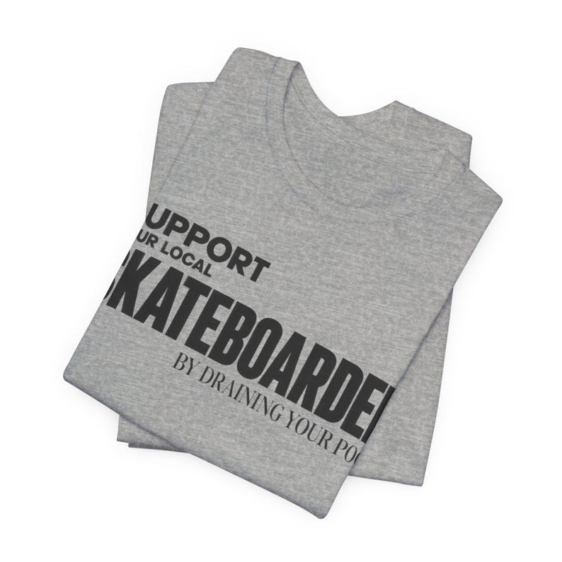 以下が含まれることがあります： SUPPORT YOUR LOCAL SKATEBOARDER BY DRAINING YOUR POOLという黒いテキストがプリントされたグレーのヘザーTシャツ。シャツは折りたたまれており、太字のサンセリフ体で描かれたグラフィックプリントが生地の前面にはっきりと表示されています。