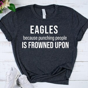 May include: Dark gray t-shirt with the word "EAGLES" in white block letters. Below the word "EAGLES" is the phrase "because punching people IS FROWNED UPON" in white. The shirt is folded on a light wood surface.