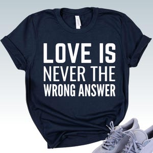 May include: Navy blue t-shirt with the text "LOVE IS NEVER THE WRONG ANSWER" in white block letters. The shirt has rolled-up sleeves and a classic crew neck design. A pair of gray sneakers is visible in the bottom right corner.