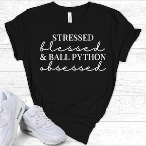 May include: Black t-shirt with the text "STRESSED blessed & BALL PYTHON obsessed" in white script. The shirt is displayed with white sneakers on a white wooden surface.