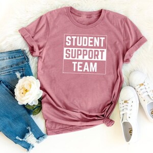 Może przedstawiać: Koszulka w kolorze malwy z nadrukiem "STUDENT SUPPORT TEAM" w kolorze białym.