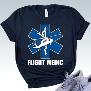 May include: A navy blue short-sleeve t-shirt featuring a white and blue Star of Life emblem with a helicopter silhouette and a serpent-entwined staff. The text FLIGHT MEDIC is printed in bold white capital letters below the central graphic design.