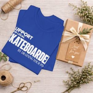以下が含まれることがあります： SUPPORT YOUR LOCAL SKATEBOARDER BY DRAINING YOUR POOLという白い文字がプリントされたロイヤルブルーのTシャツ。Tシャツは、クリーム色のリボンとJust for youと書かれたハートのタグが付いた茶色のギフトボックスの横に折りたたまれています。