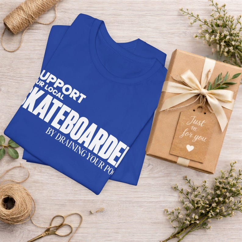 以下が含まれることがあります： SUPPORT YOUR LOCAL SKATEBOARDER BY DRAINING YOUR POOLという白い文字がプリントされたロイヤルブルーのTシャツ。Tシャツは、クリーム色のリボンとJust for youと書かれたハートのタグが付いた茶色のギフトボックスの横に折りたたまれています。